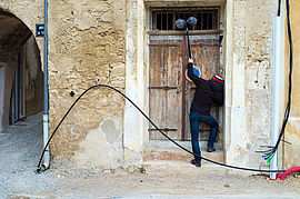 Un homme avec un blouson noir et un bonnet rouge, blanc et bleu est face à une porte en bois. Il tient une perche de prise de son.