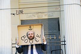 Sur l'espace public, un homme chauve, barbu et en costume tient un carton à travers lequel il passe sa tête. Sur ce carton est inscrit "Bouée" et dessiné une encre marine. Derrière lui est également inscrit sur des cartons les mots lune et étoile.